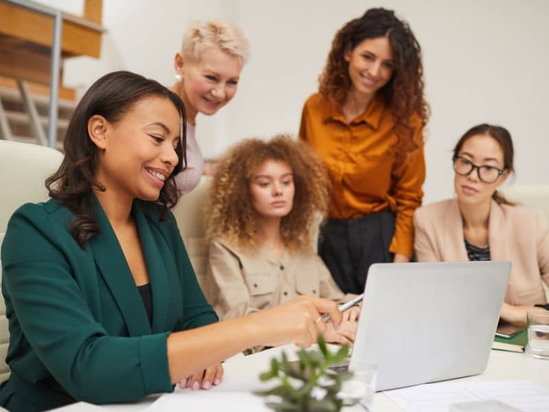 A diverse group of five women gathered around a laptop, smiling and discussing a project on a bright office table.