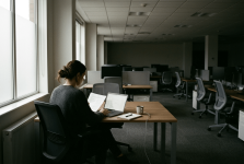 Person sitting at a wooden desk near large windows, working on a laptop with papers and a mug nearby in a quiet office.
